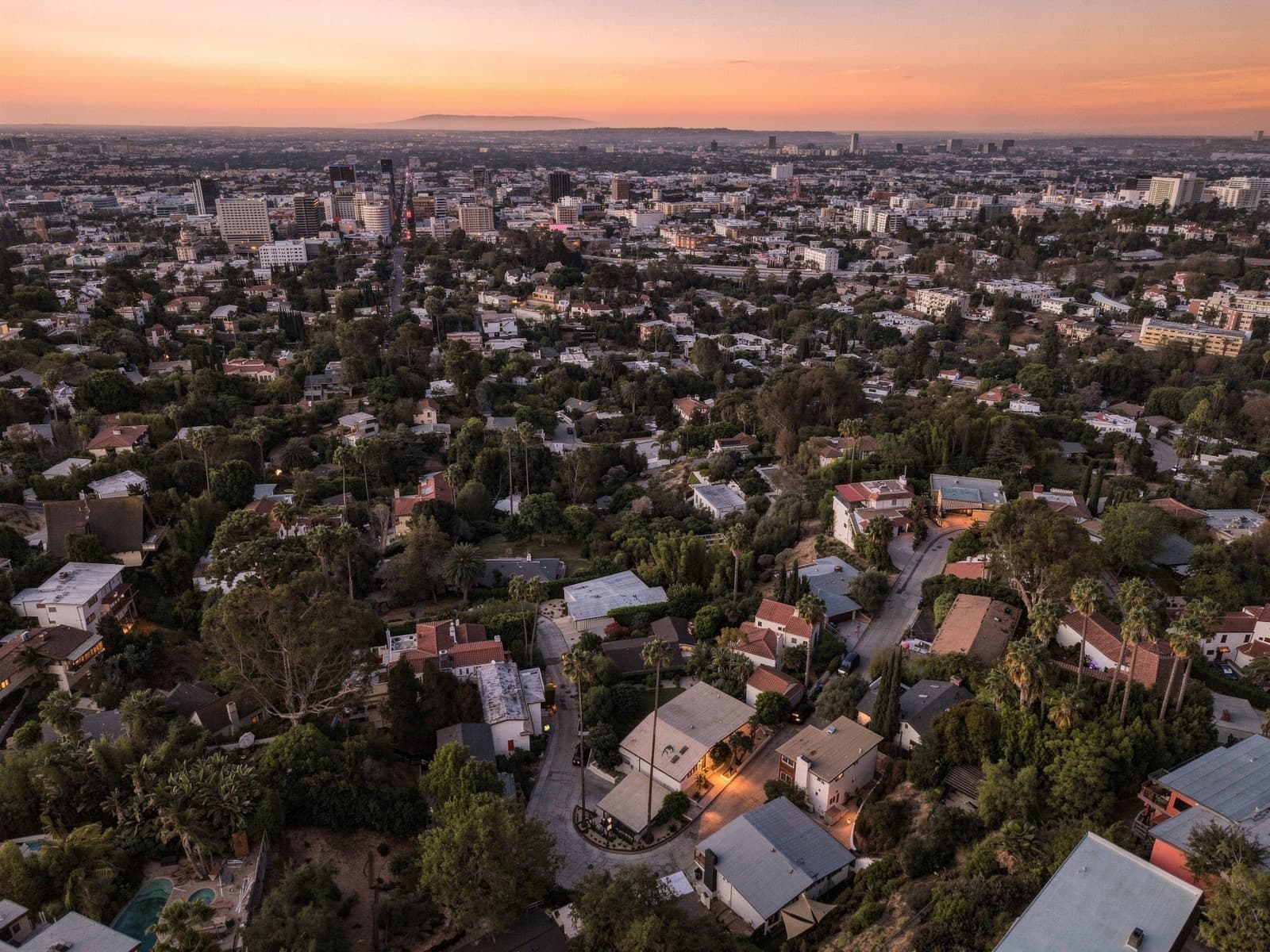 Aerial over a dense neighborhood at sunset, hillside homes and palms in front of a distant skyline under a warm orange sky