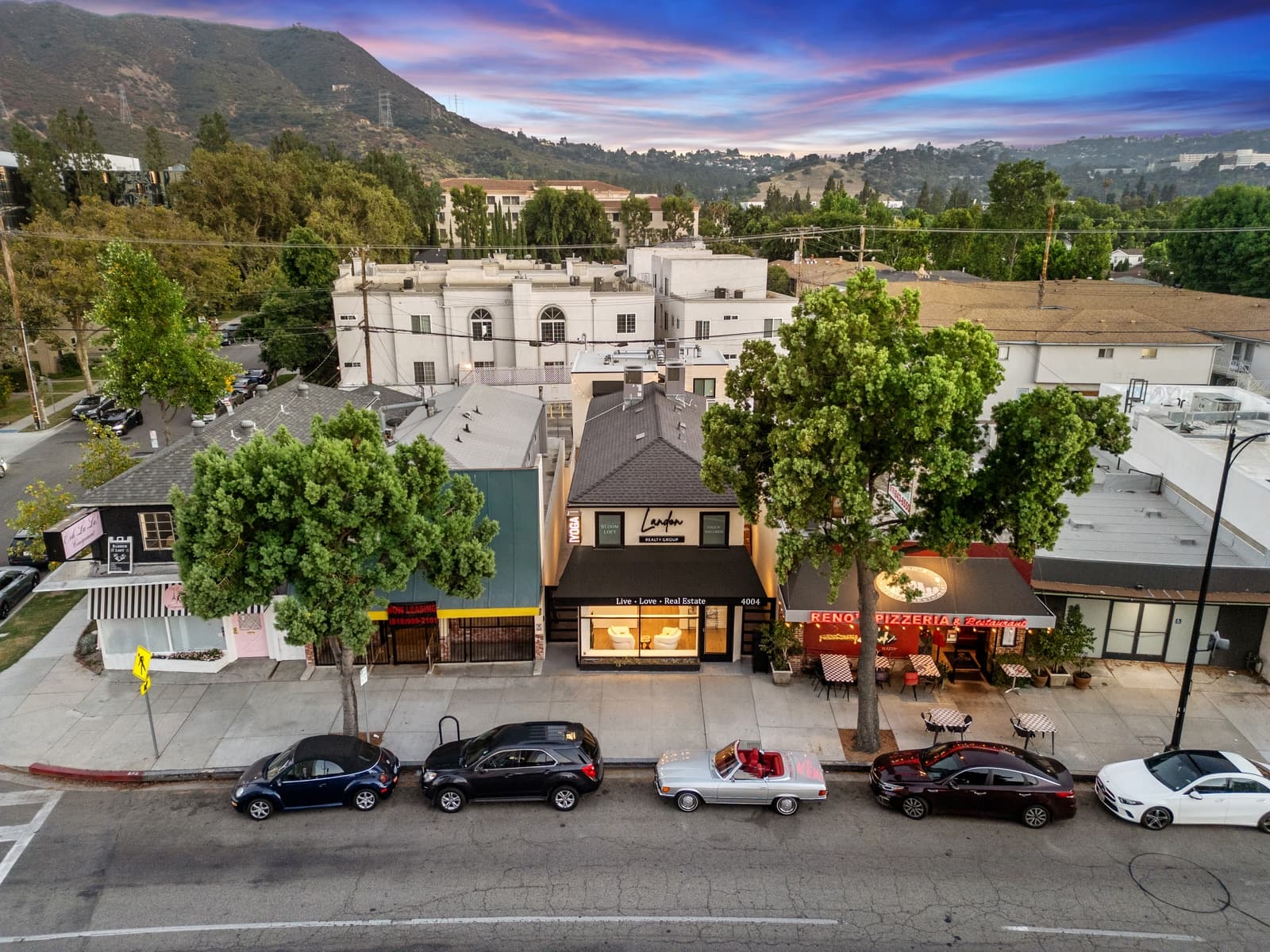 Aerial at dusk of a Burbank storefront row, a realty office and a sidewalk-dining pizzeria, with cars at the curb and hills behind