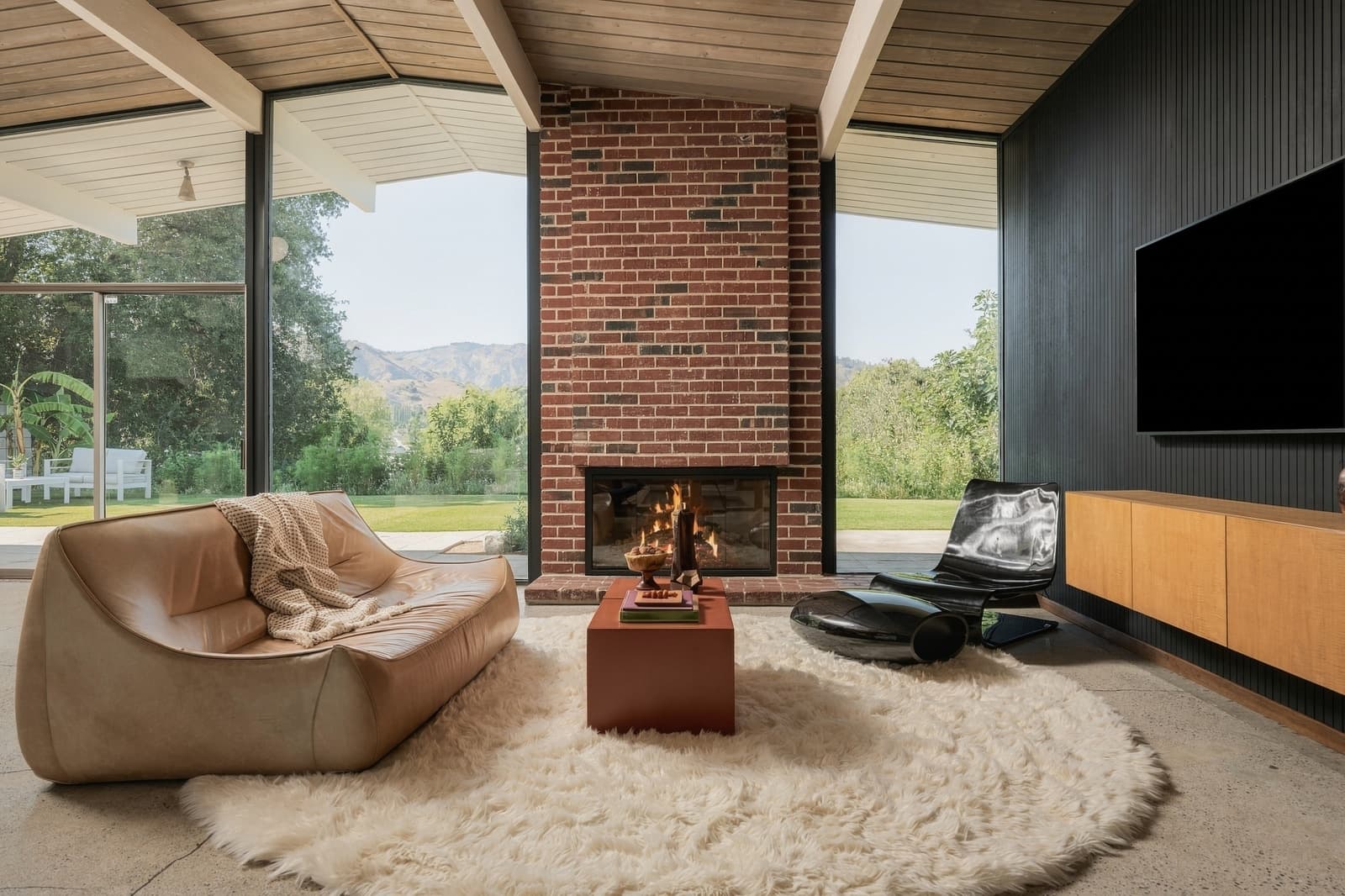 Living room with a brick fireplace and glass walls to the lawn and mountains