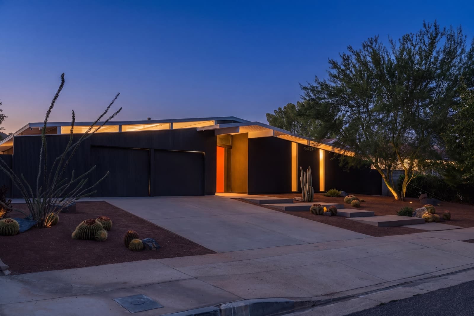 Midcentury home at twilight, warm light under the eaves and cactus in the gravel