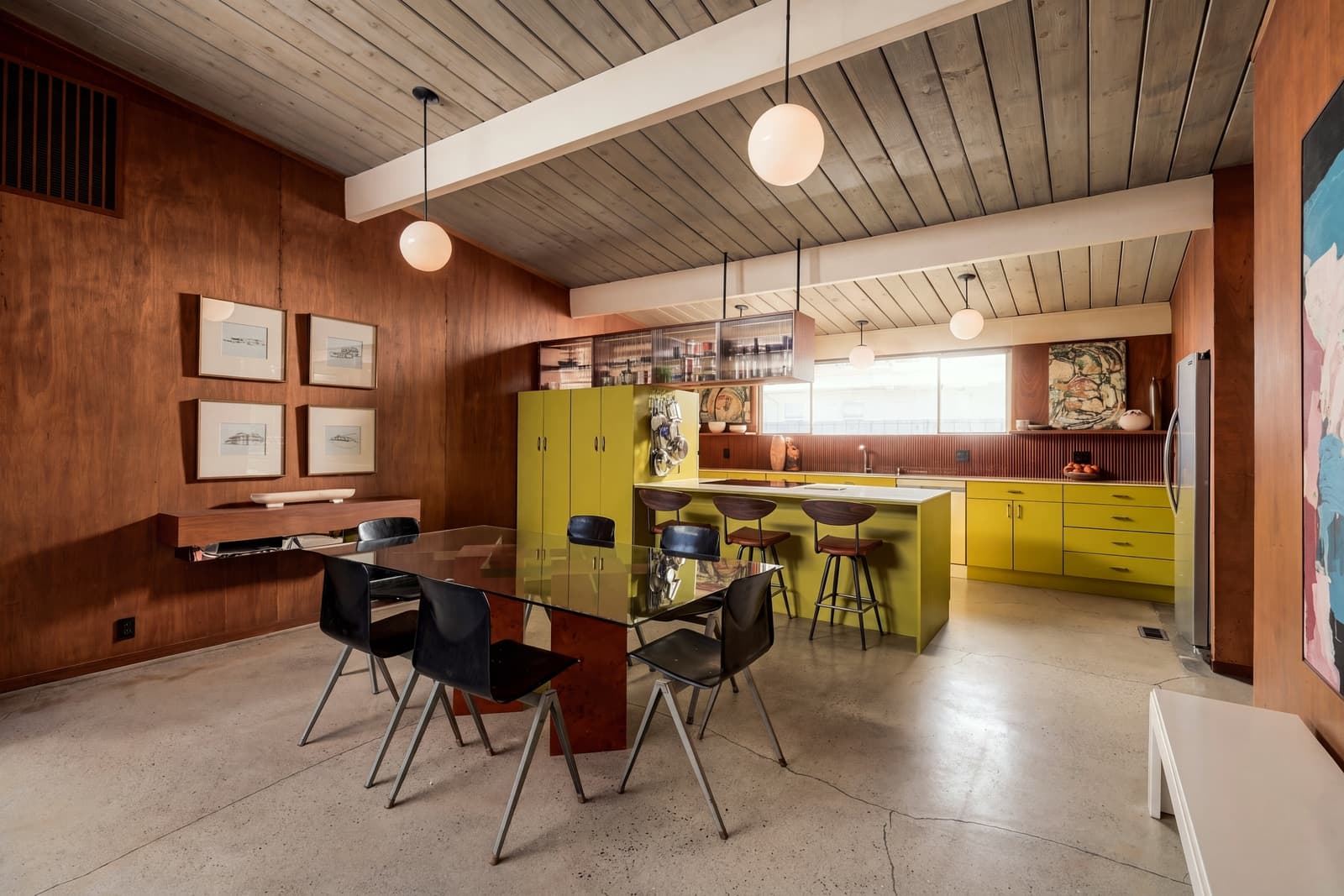 Kitchen and dining under a wood plank ceiling, yellow cabinets and globe pendants beyond the dining table