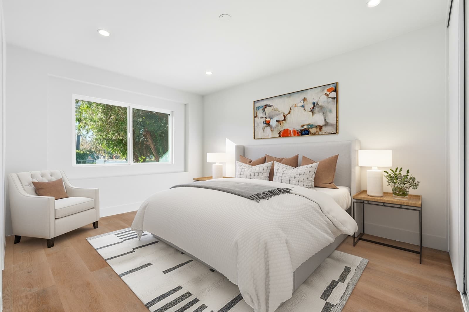 The same bedroom virtually staged with an upholstered bed, layered white bedding, lamps on two nightstands, a cream armchair and a striped rug