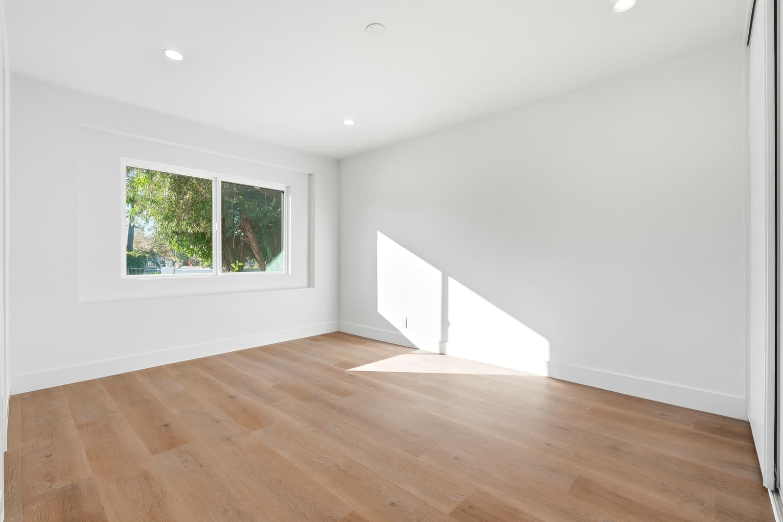 Empty bedroom with a wide sliding window looking onto leafy trees, white walls and light wood floors