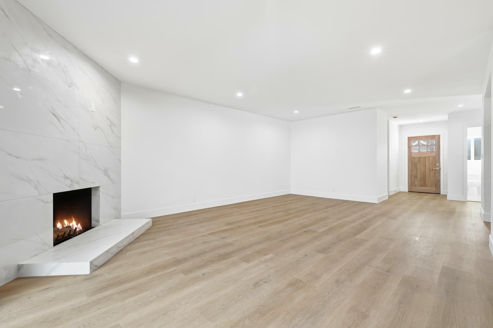 Empty living room with a floor-to-ceiling marble-tile fireplace, a lit fire and pale wood floors