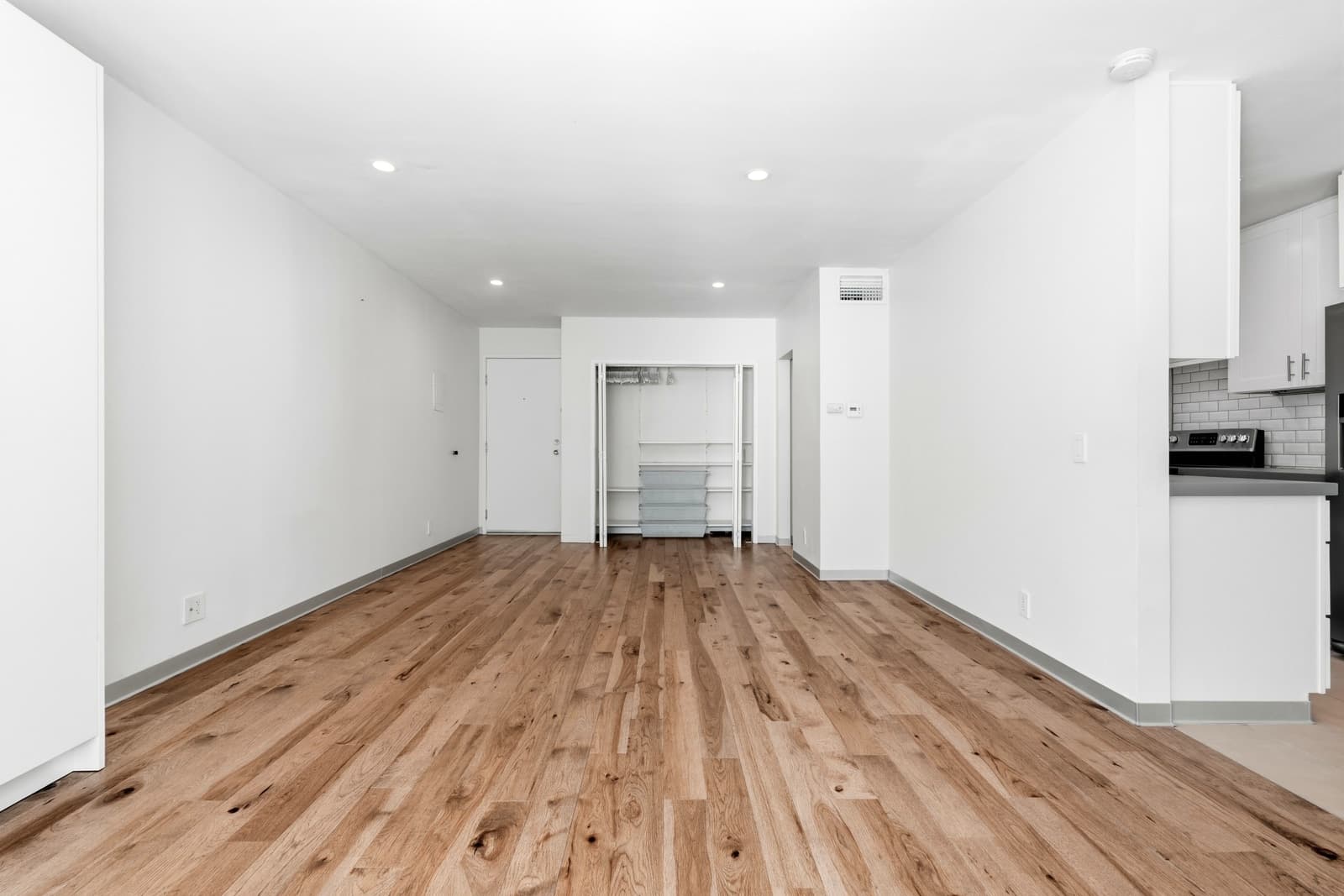 Empty open living room beside a white kitchen, with wide-plank floors and an open closet by the entry door