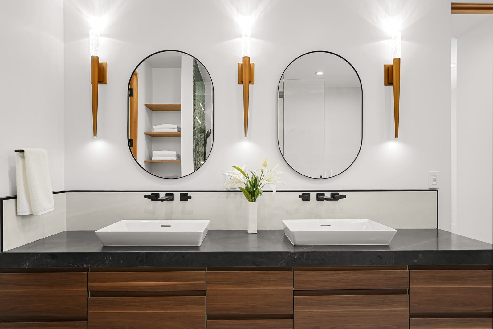 Double vanity with oval mirrors and brass sconces on a black stone top