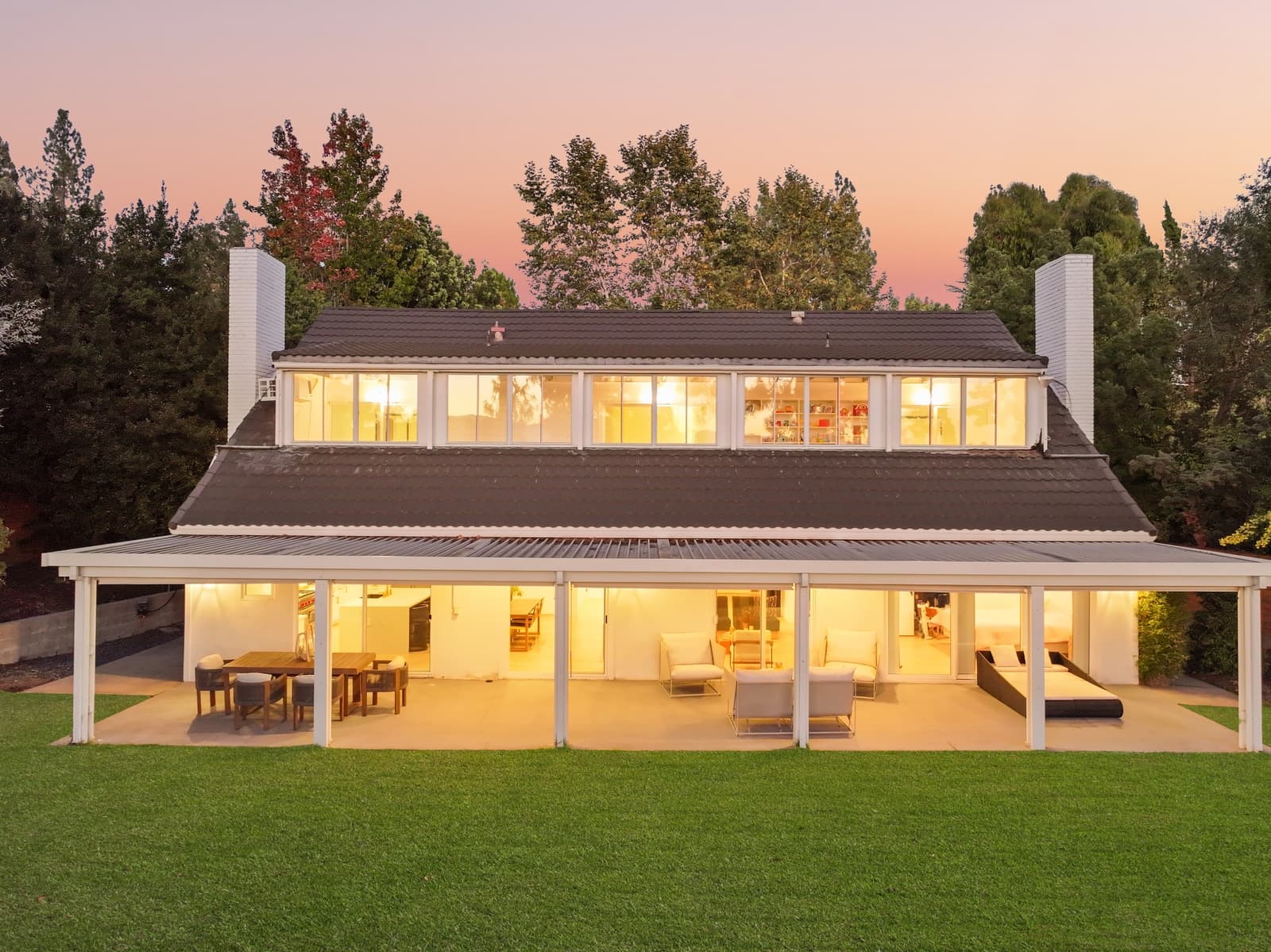 Two-story home at dusk seen across the back lawn, every window glowing above a covered patio