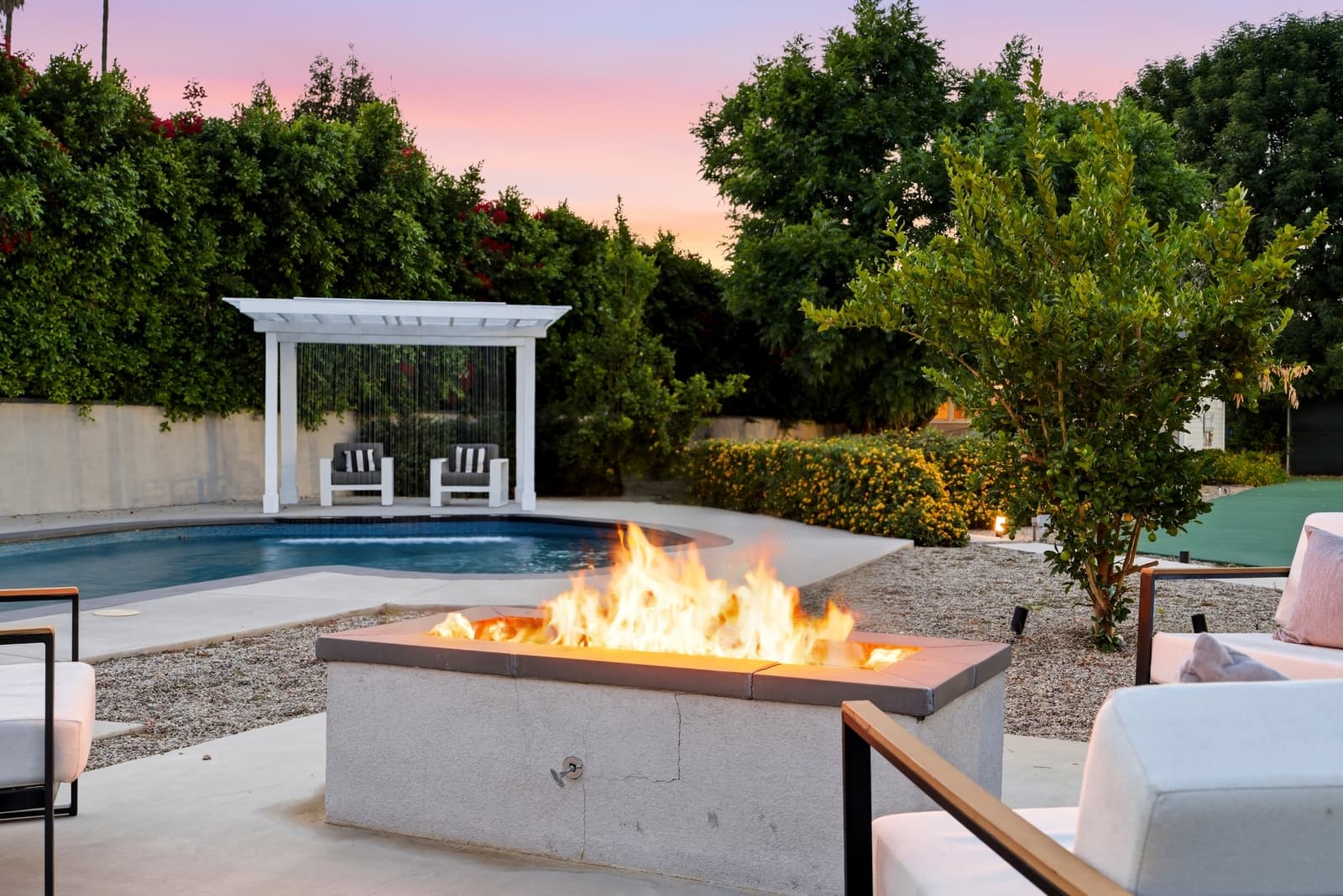 Fire table burning by the pool at dusk in Woodland Hills, a white pergola and lounge chairs behind